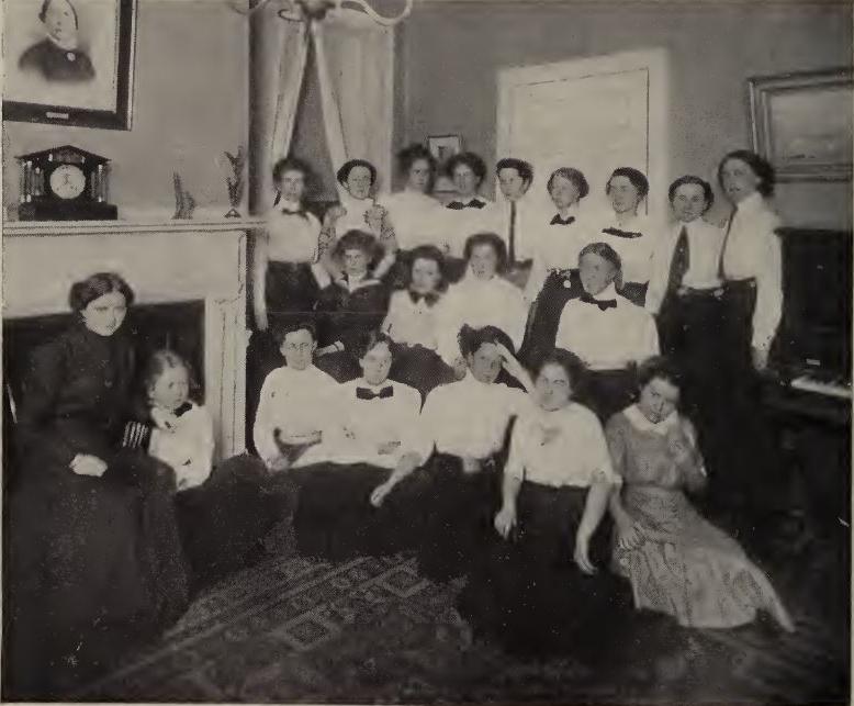 20个人的黑白照片. All three rows. 大多数人都穿着白色上衣和深色裤子. 在照片的右侧，一个穿着深色连衣裙的女人坐在壁炉前.